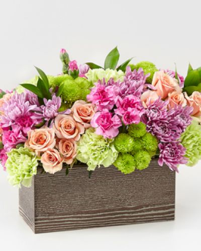 Capture the beauty of the seasons in bloom with our Simple Charm Bouquet. Gorgeous blooms such as peach spray roses, green trick dianthus, pink mini carnations and lavender cushion pompons fill a weathered wooden box with freshness.